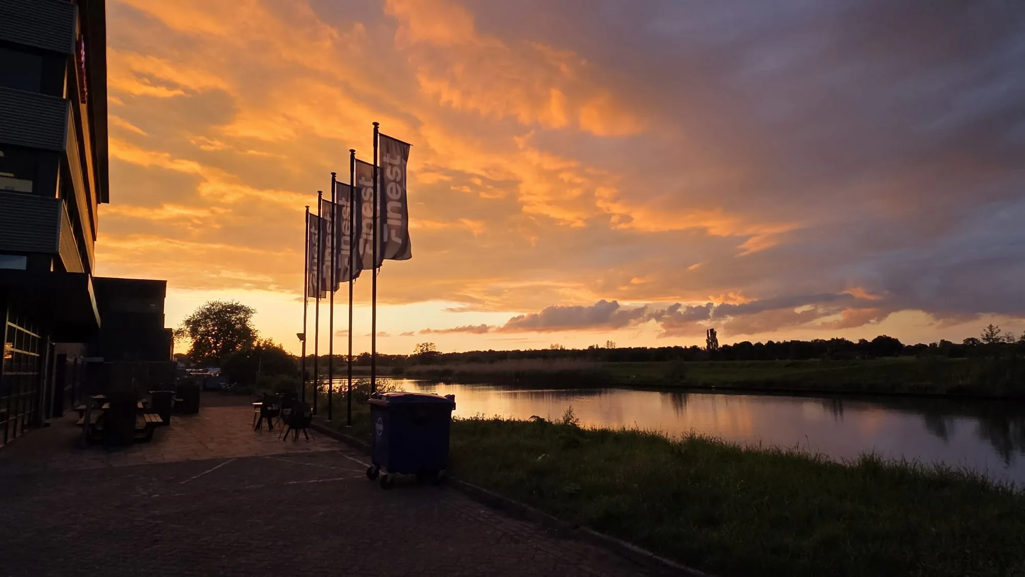 Kantoorpand aan het water met een rij vlaggen op de kade, onder een oranje avondlucht